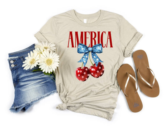 BELLA CHERRY AMERICA CREAM 4TH OF JULY GRAPHIC TEE featuring red star-spangled cherries with a blue bow and bold 'AMERICA' text, styled with denim shorts and flip-flops.