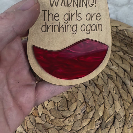 Hand holding ornament with 'Warning! The girls are drinking again'
